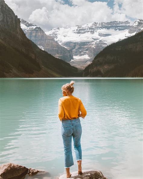 A Woman Standing On The Edge Of A Lake Looking At Mountains And Snow