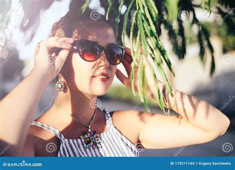 Portrait Of An Attractive Girl In Sunglasses Hiding Behind Tree Branches On A Bright Sunny Day