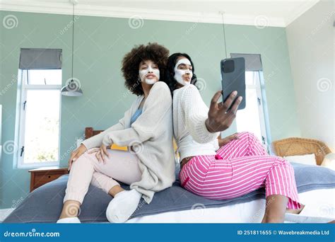 Biracial Friends With Cream On Faces Posing And Taking Selfie Over Smartphone While Sitting On