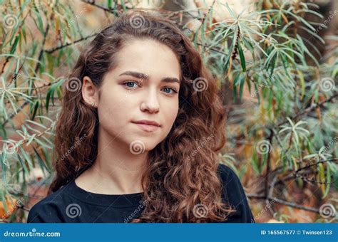Portrait Of A Girl With Brown Curly Hair On A Background Of Green Bush