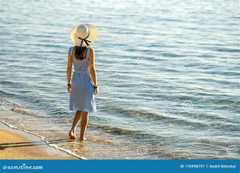 Young Woman In Straw Hat And A Dress Standing Alone On Empty Sand Beach