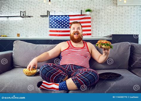 Thick Funny Man Eating A Burger Sitting On The Couch Against The Stock