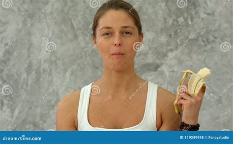 Young Woman Holding Banana Eating It Looking At Camera Stock Photo