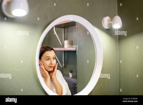 woman in bathrobe making facial massage looking into the round mirror in the bathroom stock