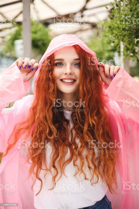 Beautiful Lady With Redhead Curly Hair Standing In Pink Raincoat And Happily Looking In Camera