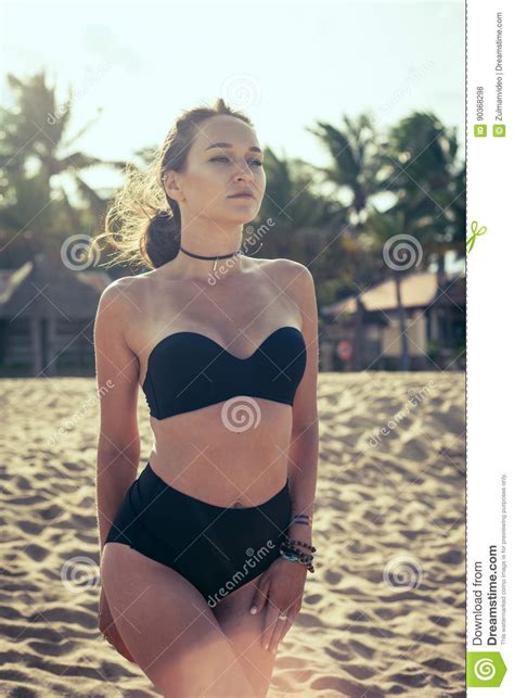 Beautiful Woman With Tan Skin In A Black Swimsuit Standing On The Sand