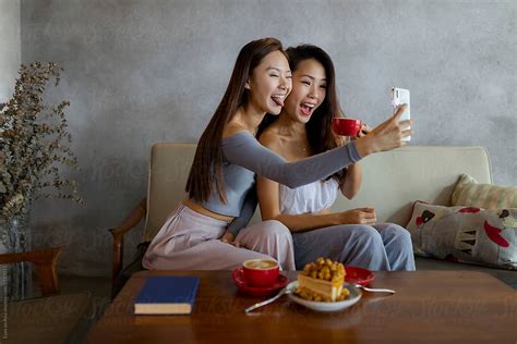 Two Asian Female Friends Taking Selfies Together With Coffee And Cake Sitting On A Couch Del