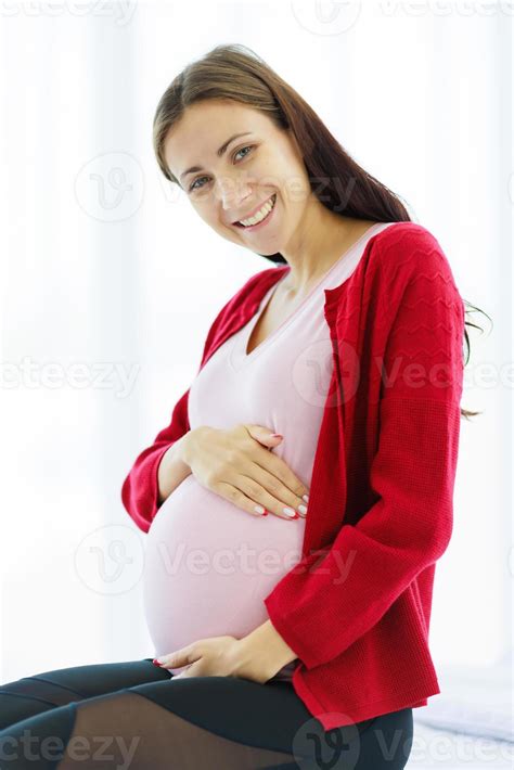 pregnant woman sits on the bed in her bedroom caressing her belly with a happy smiling face