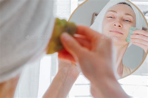 woman in bathrobe and a towel on her head looking in the mirror and making face massage with