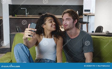 Happy Interracial Couple Making Selfie Video Call Via Smartphone While Relaxing On Sofa At Home