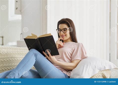 A Girl In Glasses Reads Book Sitting On The Couch In The Room Stock