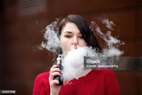 Vape 십대 겨울에 거리에서 현대 갈색 배경 맞은 편에 전자 담배를 흡연 빨간 스웨터에 젊은 예쁜 흰색 백인 소녀 나쁜 습관 18 19세에 대한 스톡 사진 및 기타 이미지