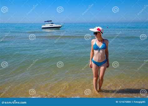 A Blond Girl Wearing Sunglasses And A White Hat Is Standing On The Beach Opposite The Yacht