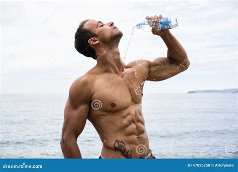 A Shirtless Man Standing On The Beach With A Bottle In His Hand And