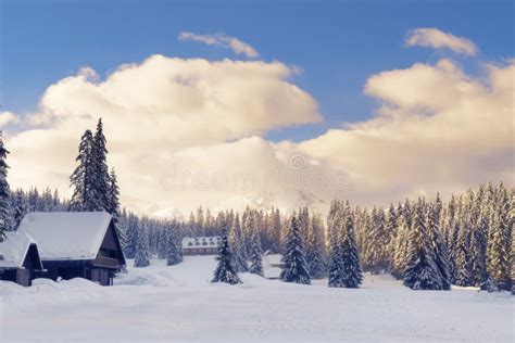 Wooden Cabins In Winter Landscape At Sunset With Sunlit Mountains In