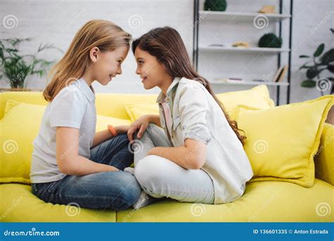 Happy And Overjoyed Girls Sit On Sofa Face To Face They Look Into Eyes