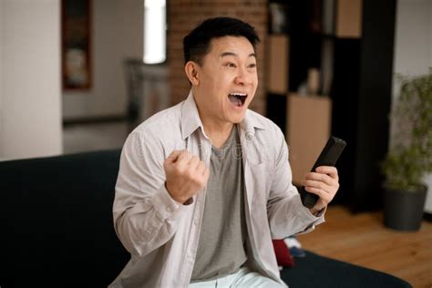 Emotional Mature Asian Man Watching Sport On Tv Celebrating Success And Screaming Holding Tv