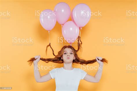 Redhead Girl With Balloons Tied To Hair Blowing Cheeks And Looking At Camera Isolated On Yellow