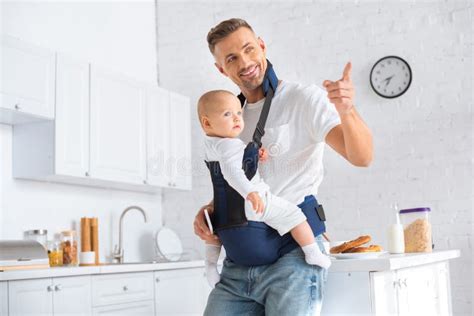 Cheerful Father Holding Infant Daughter In Baby Carrier And Pointing