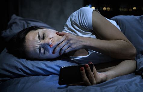 Young Sleepy Female Using Mobile Phone Yawning Late At Night Lying In