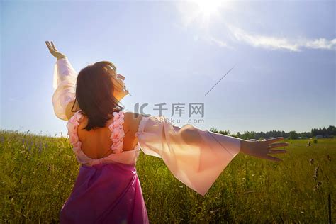 在鲜花盛开和蓝天背景下，身着郁郁葱葱的粉红色舞会礼服的漂亮女孩在绿色的田野里。高清摄影大图 千库网
