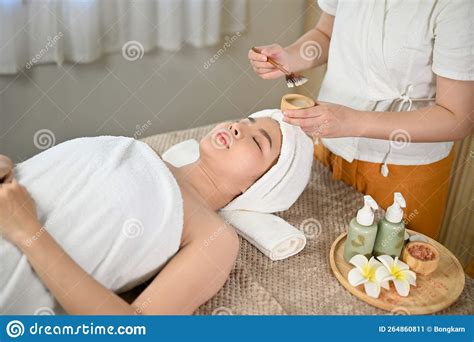 relaxed asian woman lying on massage table with eyes closed getting facial treatment stock