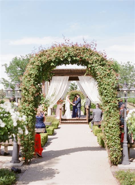A Couple Getting Married Under An Arch Covered In Flowers And Greenery