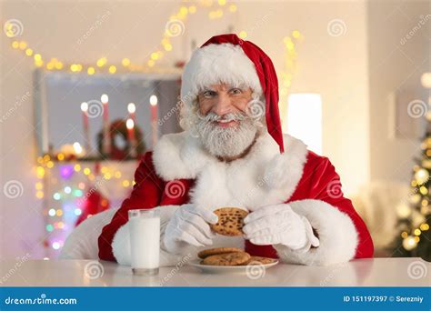 Santa Claus Eating Cookies And Drinking Milk At Table In Room Decorated