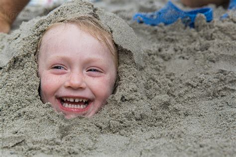 Happy And Smiling Child Buried In Sand At Beach By Stocksy