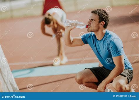 Sporty Man Drinking Water While Young Woman Doing Exercise In Th Stock