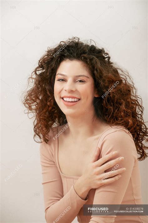 cheerful dreamy female with curly hair and in bodysuit sitting on floor against white wall in