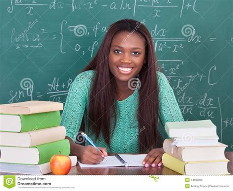 Confident Female Teacher Writing In Book At Classroom Desk Stock Image