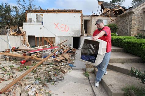 A Man Carrying A Large Box Full Of Debris In Front Of A Destroyed House