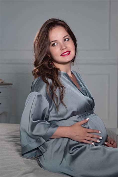 Happy Pregnant Woman Sitting On Bed And Touching Her Belly At Home