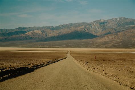 A Long Dirt Road Leading Towards The Rocky Mountains In An Arid