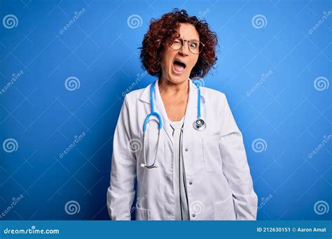 middle age curly hair doctor woman wearing coat and stethoscope over blue background winking