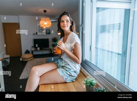 Smiling Young Woman Sitting On Kitchen Counter With A Drink Stock Photo