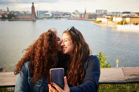 Smiling Lesbian Couple Kissing While Taking Selfie With Mobile Phone Against Cityscape Stock Photo