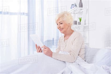 Side View Of Happy Senior Woman With Tablet Lying In Bed At Home