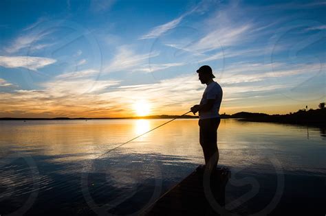 Silhouette Of A Young Man Fishing At A Lake In Portugal At Sunset With