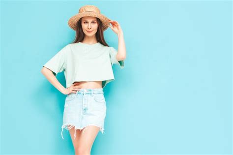 스튜디오의 파란색 벽 근처에서 포즈를 취한 트렌디한 여름 청바지 치마를 입은 평온한 여성의 초상화 실내에서 즐거운 시간을 보내는 긍정적인 모델 쾌활하고 행복한 모자 무료 사진