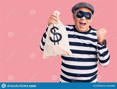 senior handsome man wearing burglar mask holding money bag screaming proud celebrating victory