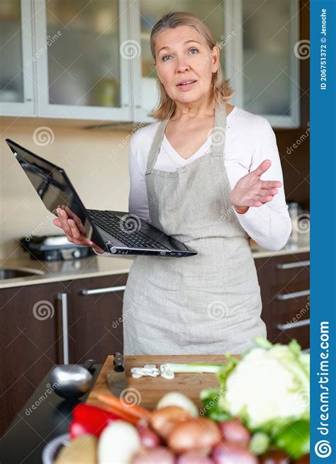 Mature Woman In The Kitchen Preparing Food And Holding Laptop Stock