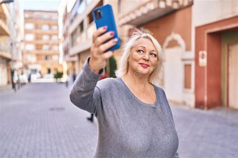 middle age blonde woman smiling confident making selfie by the smartphone at street stock image