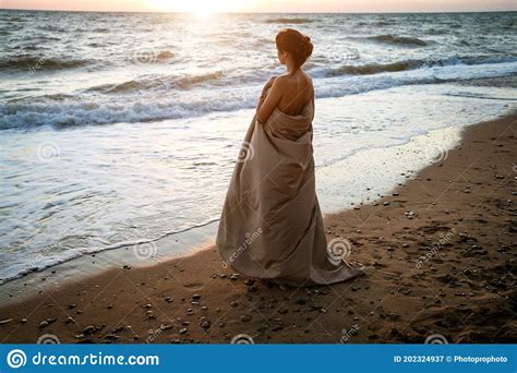 A Beautiful Woman Stands On The Seashore At Sunset Wrapped In A