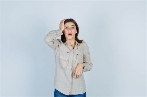 Молодая девушка с рукой на голове держит рот открытым в бежевой рубашке джинсах и выглядит