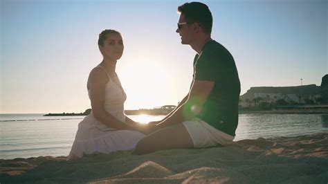 A Young Couple Is Sitting On The Sandy Seashore Smiling Holding Hands