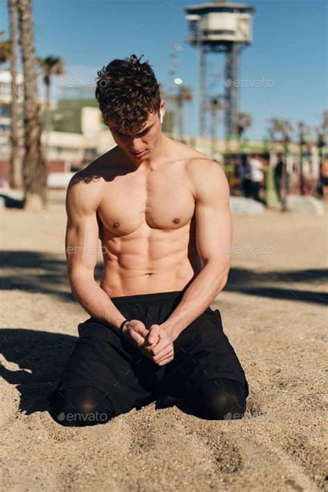handsome sporty guy with naked torso resting on sand after workout on hot summer day on beach