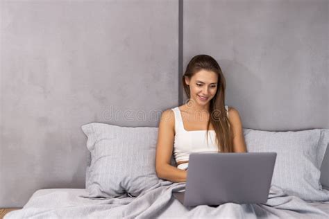 smiling woman catching up on her social media as she relaxes in bed with a laptop on a lazy day