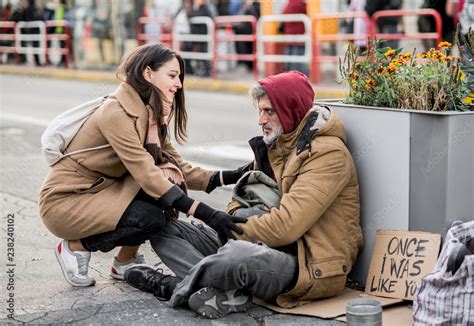 Young Woman Giving Money To Homeless Beggar Man Sitting In City Stock